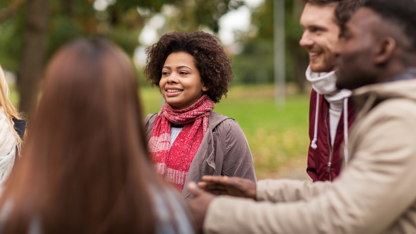 Four people in their 20's in a green space. Black woman is smiling and in focus, two men are out of focus and we can see the back of another woman's head. 